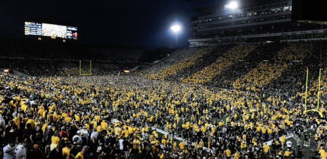 Fans rush the field after Iowa defeats Penn State, 23-20, amid a backdrop of black and gold striped stadium sections.