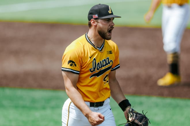 Iowa pitcher Will Christopherson yells victoriously as he walks off the mound. Iowa is wearing gold jerseys with Iowa written in script, black caps with a script "I", and white pants.